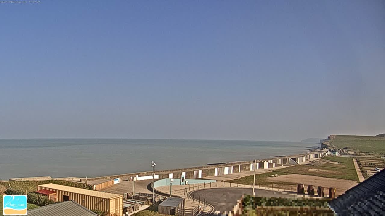 A calm, clear day overlooks a coastal town with a seawall, beach huts, and cliffs in the distance.