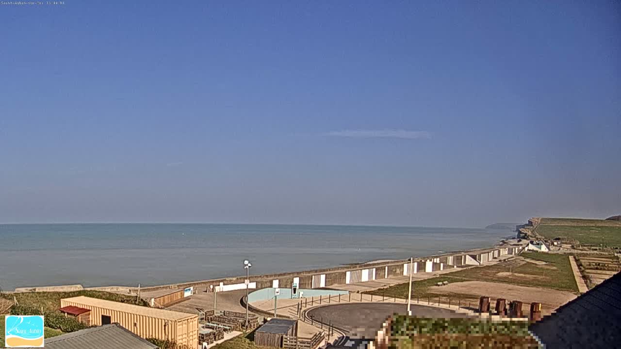 A coastal scene under a clear blue sky shows a calm sea, a seawall with beach huts, and a grassy area leading up to a cliff.