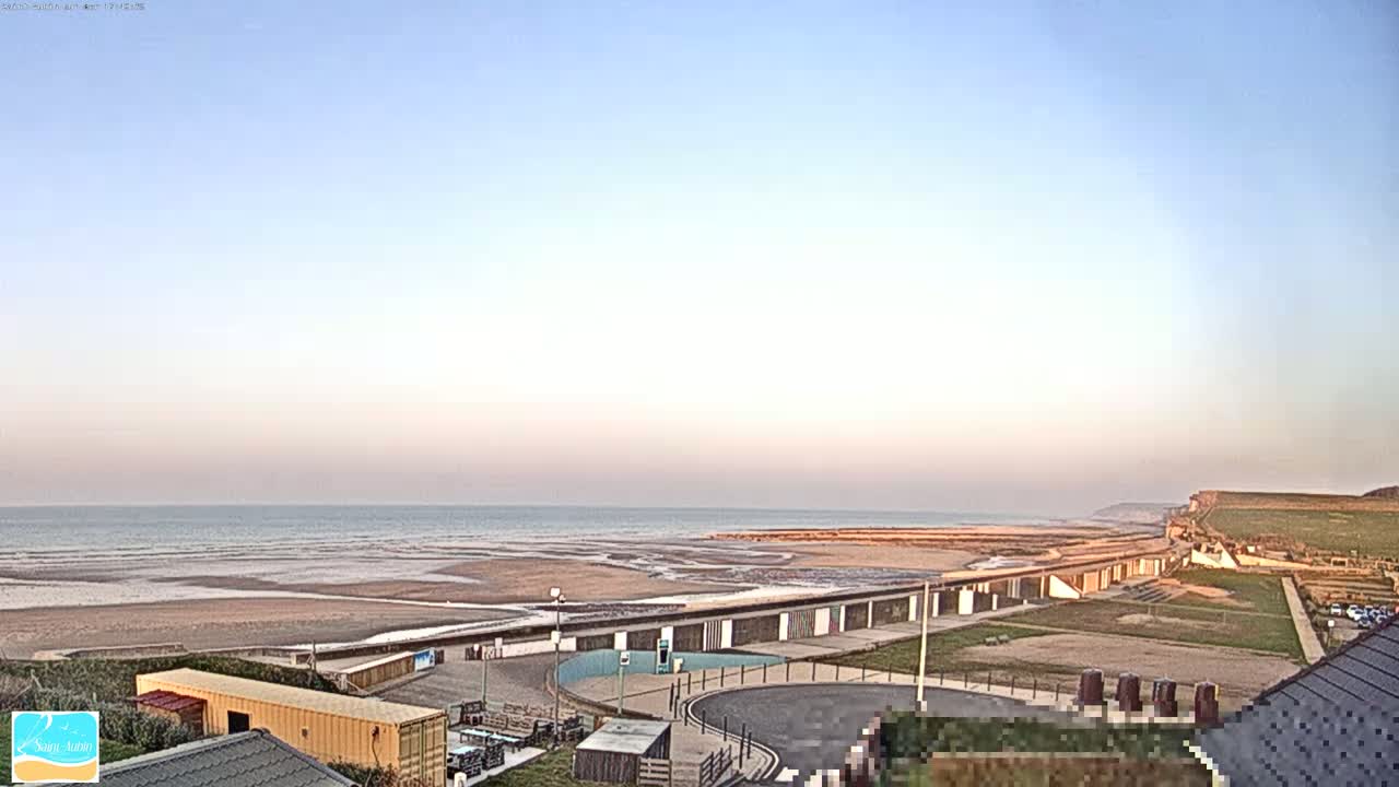 A sandy beach stretches along a calm ocean under a clear, pale sky, with beach huts and a grassy cliff visible in the foreground.