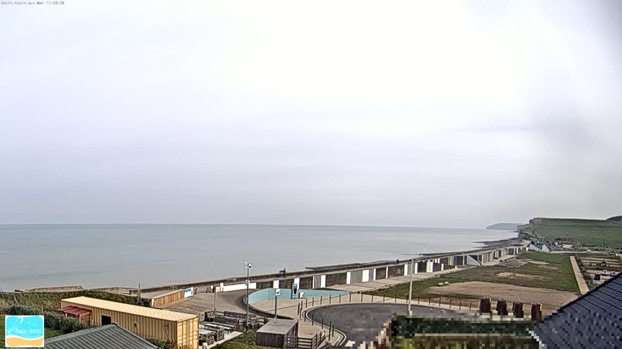 A calm, overcast day reveals a seaside view of a beach with a long row of beach huts, a paved area, and a grassy cliff in the distance.