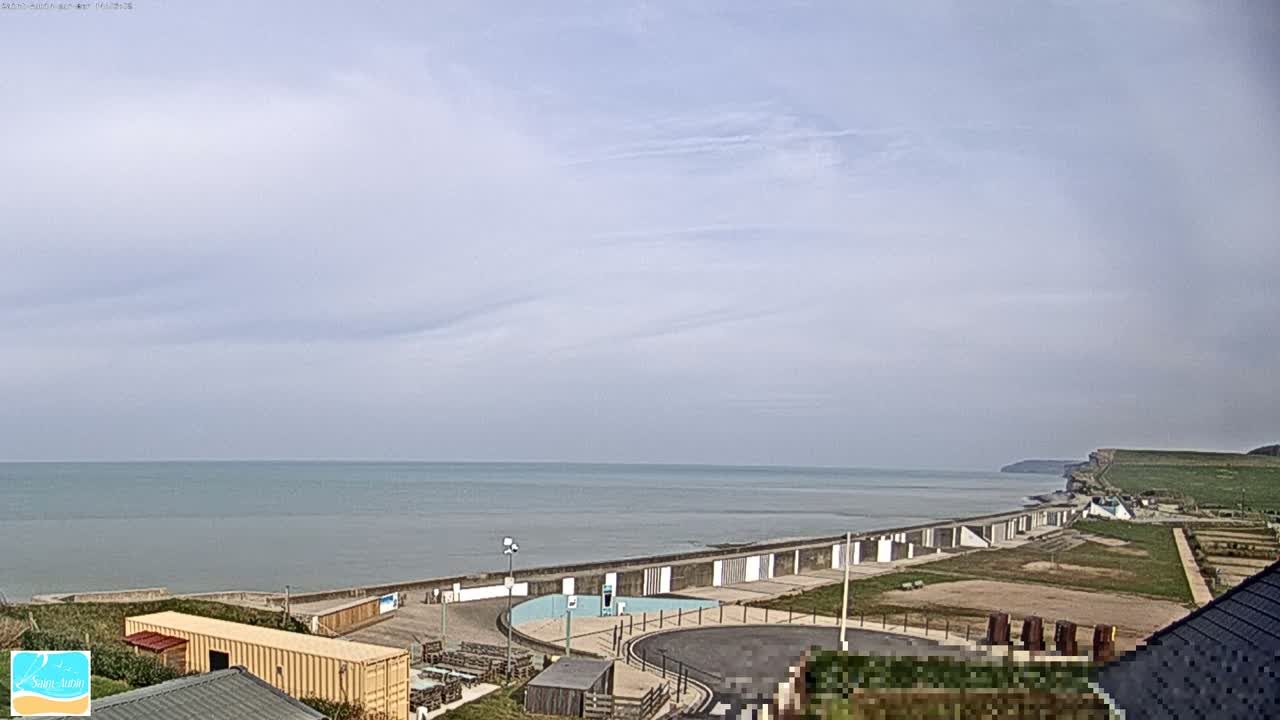 A coastal scene under a mostly cloudy sky shows a calm sea meeting a seawall and beachfront area with buildings and grassy areas, extending to cliffs in the distance.