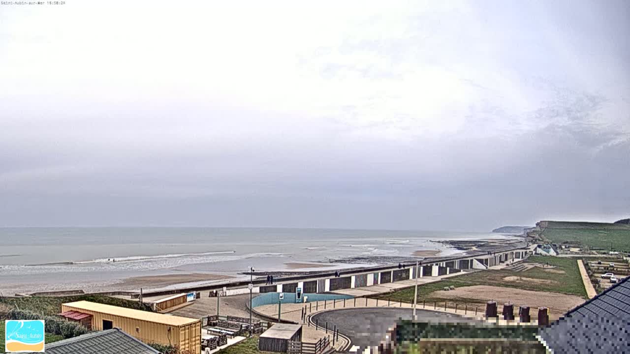A beach with a row of beach huts is visible under an overcast sky, with a calm sea and cliffs in the distance.