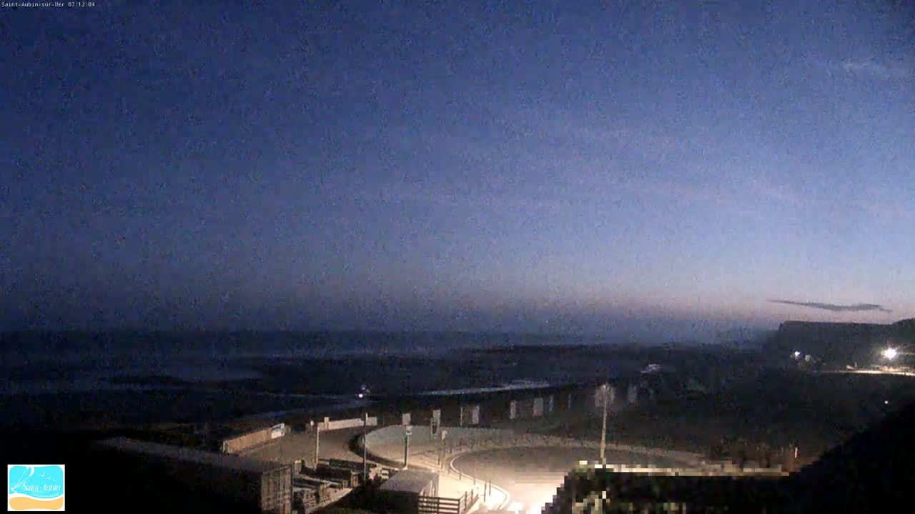 A dark, pre-dawn coastal scene shows a calm ocean under a twilight sky, with a curved roadway and buildings visible near the shore.
