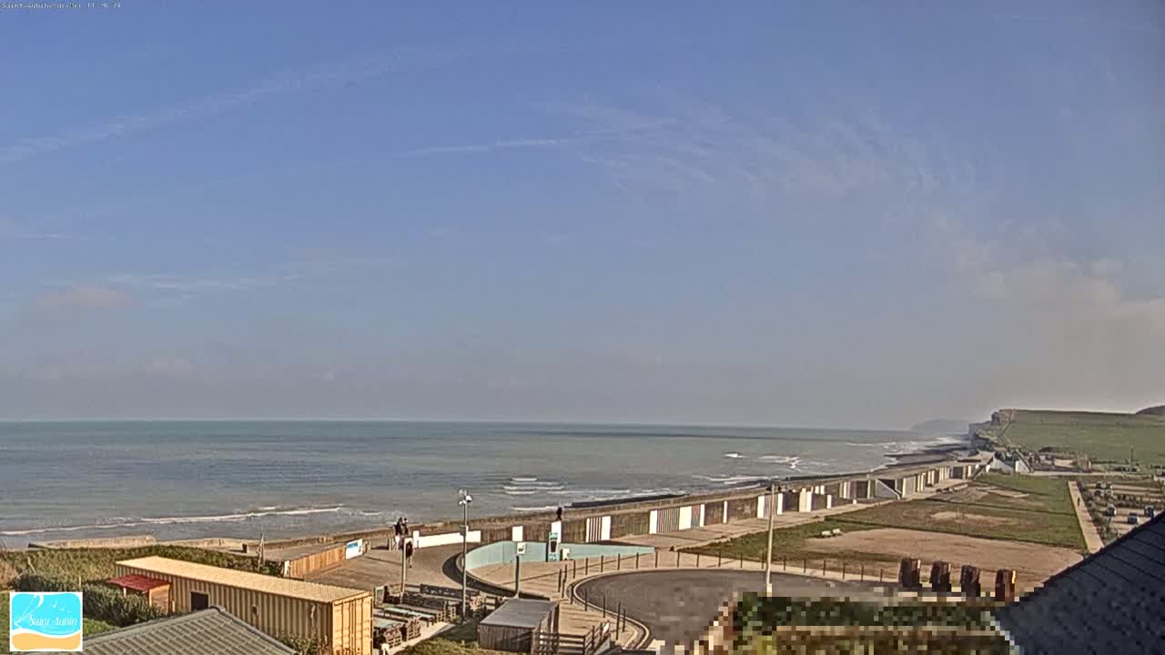 A mostly clear day shows a beach with a sea wall, a parking area, and some buildings, all overlooking a calm ocean with small waves and distant cliffs.