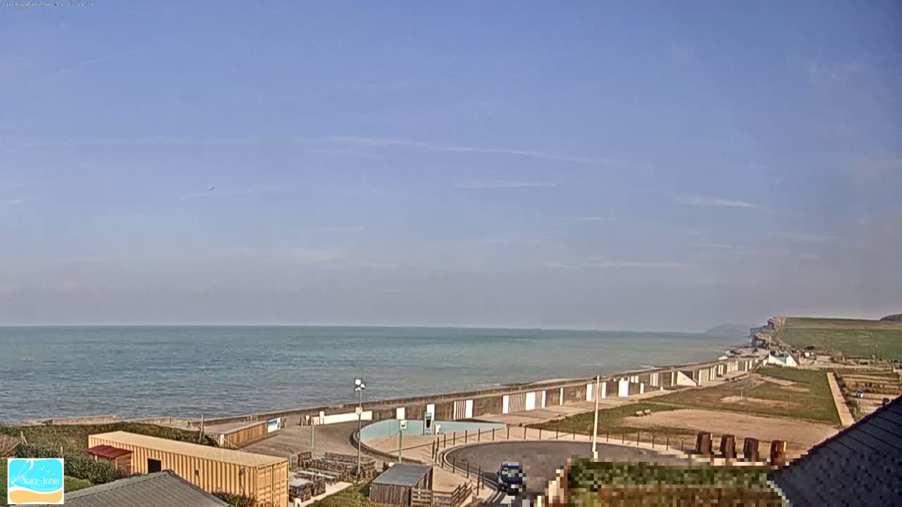 A calm ocean meets a coastline with a seawall, beach huts, and grassy areas under a clear, blue sky.
