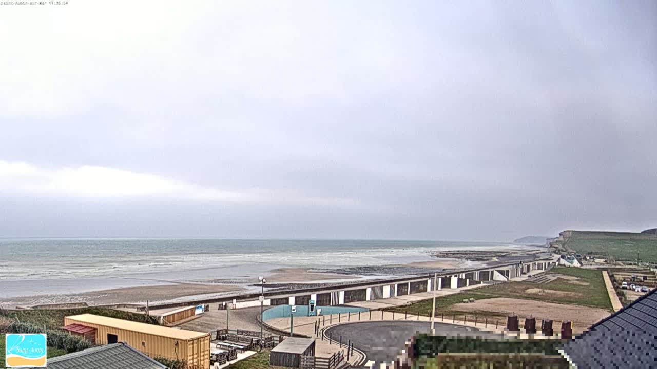 A beach with calm waves under an overcast sky features a long row of beach huts and a cliff in the distance.