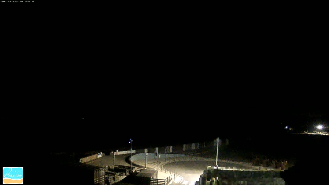 A nighttime, dimly lit, high-angle view of a coastal road curving around a small, paved area.