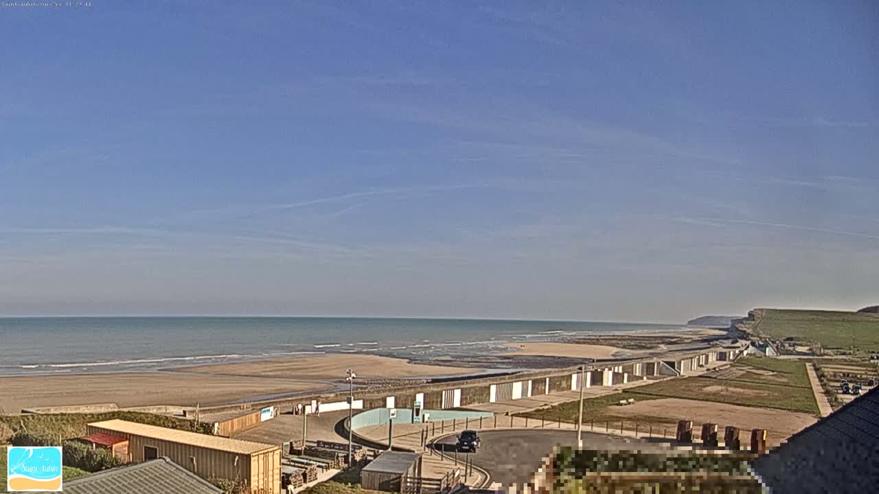 A mostly sunny day shows a long sandy beach with calm waves and a seawall extending along the coast, near a cliff, with a small parking lot and buildings visible in the foreground.