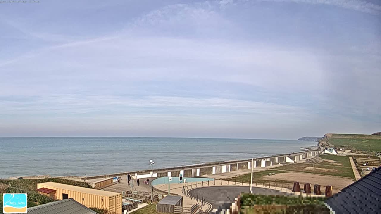 A calm ocean stretches to the horizon under a mostly clear sky, with a seawall and beachfront area visible in the foreground.