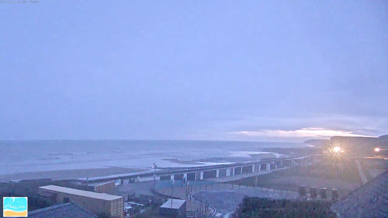 A calm, overcast ocean beach scene is shown with a long structure situated between the beach and a road, under a pale sky.