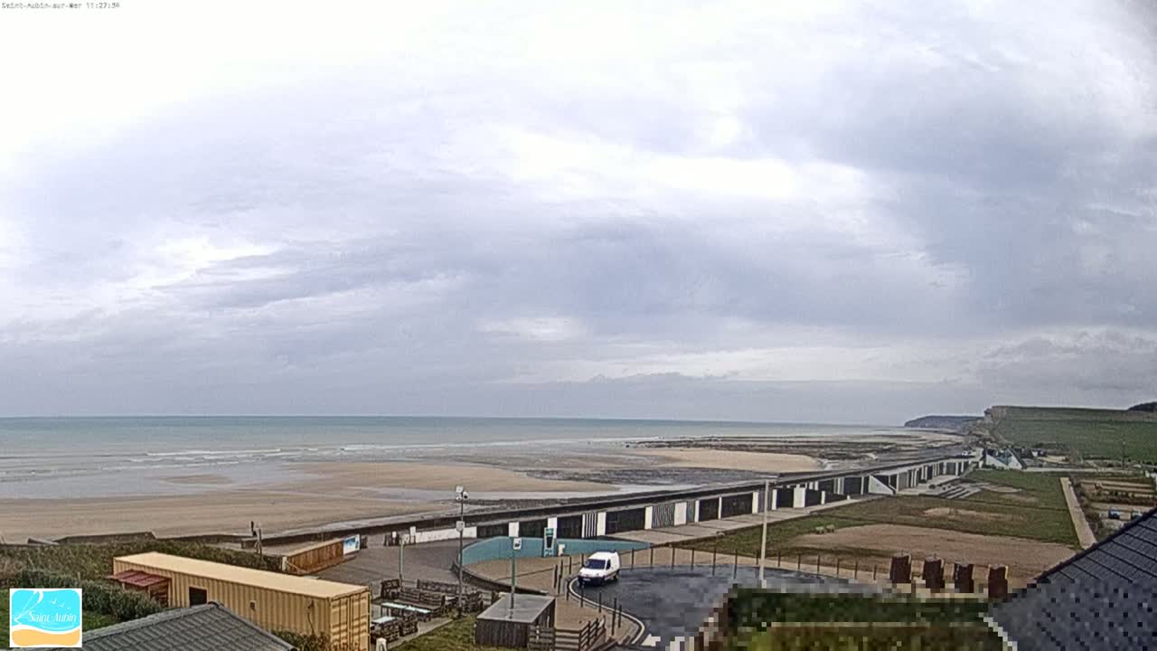 A wide view of a beach with overcast skies, showing calm waves, sandy shores, buildings, and a van parked near a seawall.