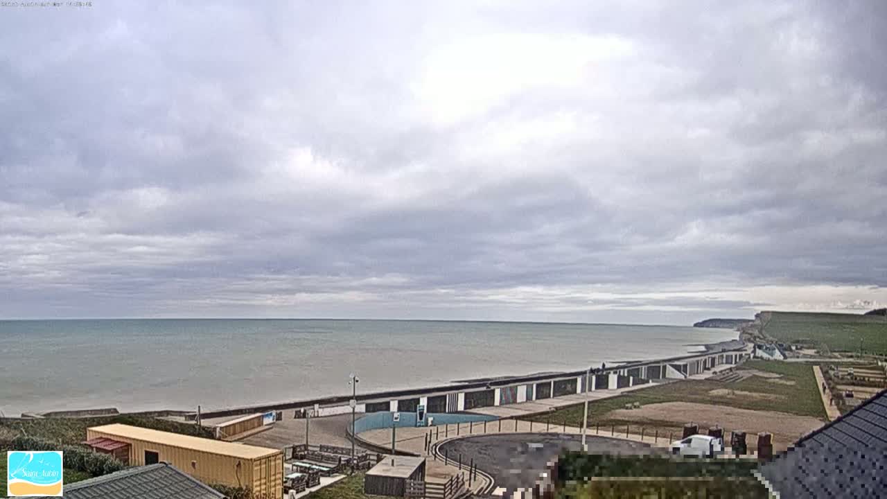 A calm sea stretches to the horizon under a mostly cloudy sky, with a beachfront area featuring buildings and a parking lot visible in the foreground.