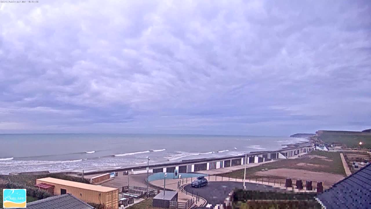 A mostly cloudy sky overlooks a beach with small waves, a seawall, and a few buildings.
