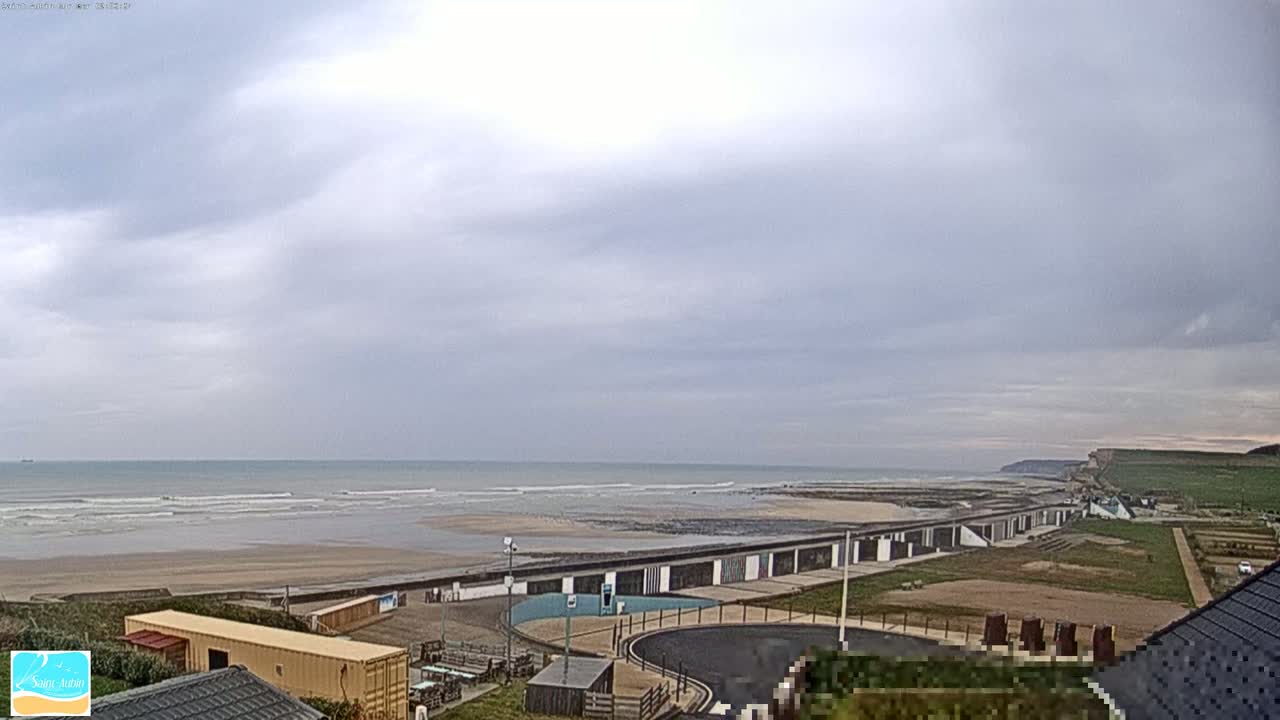 A beach with small waves under an overcast sky is viewed from a high vantage point, showing a long line of beach huts and a paved area leading to the shore.