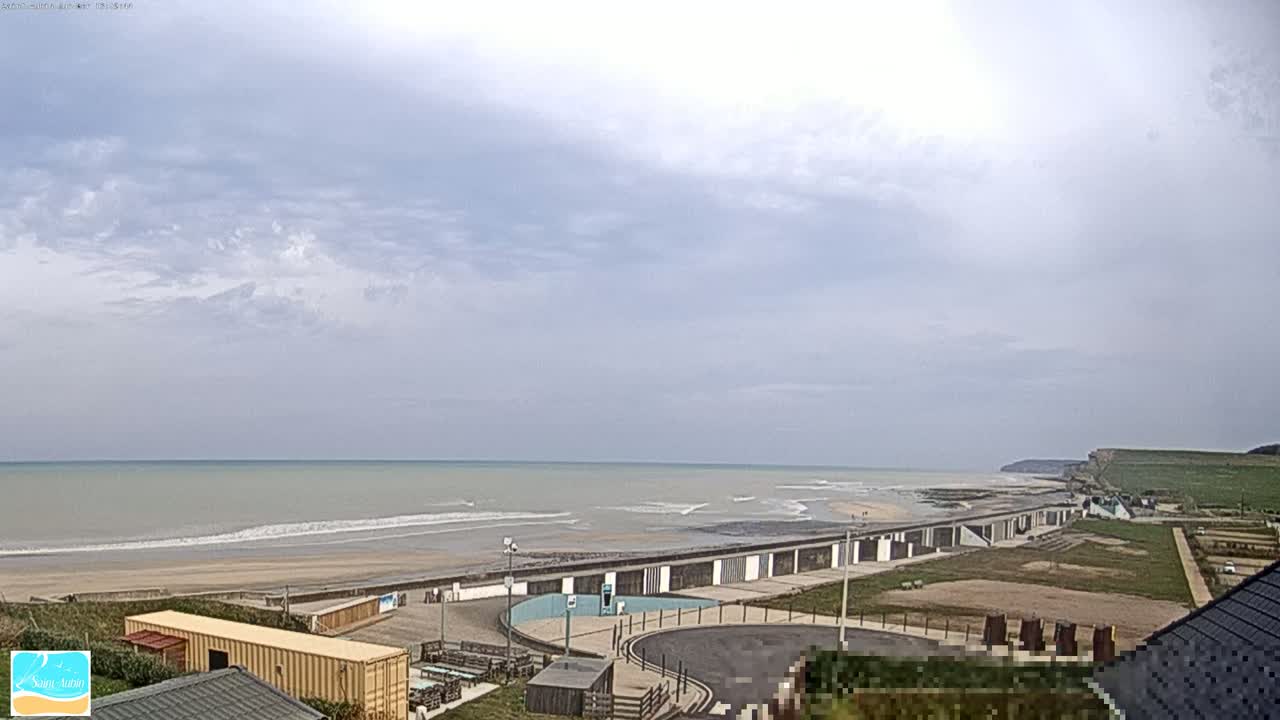 A calm, overcast day shows a view of a beach with small waves, a seawall, and buildings, all overlooking a grassy area and cliffs in the distance.