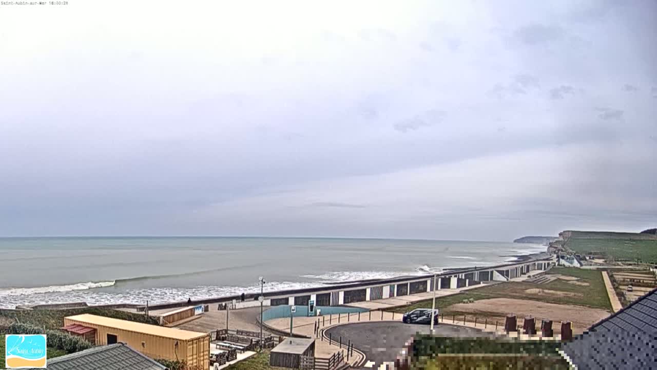 A mostly cloudy day reveals a calm ocean with small waves lapping a shoreline featuring a long row of beach huts and a parking lot, all backed by grassy hills.