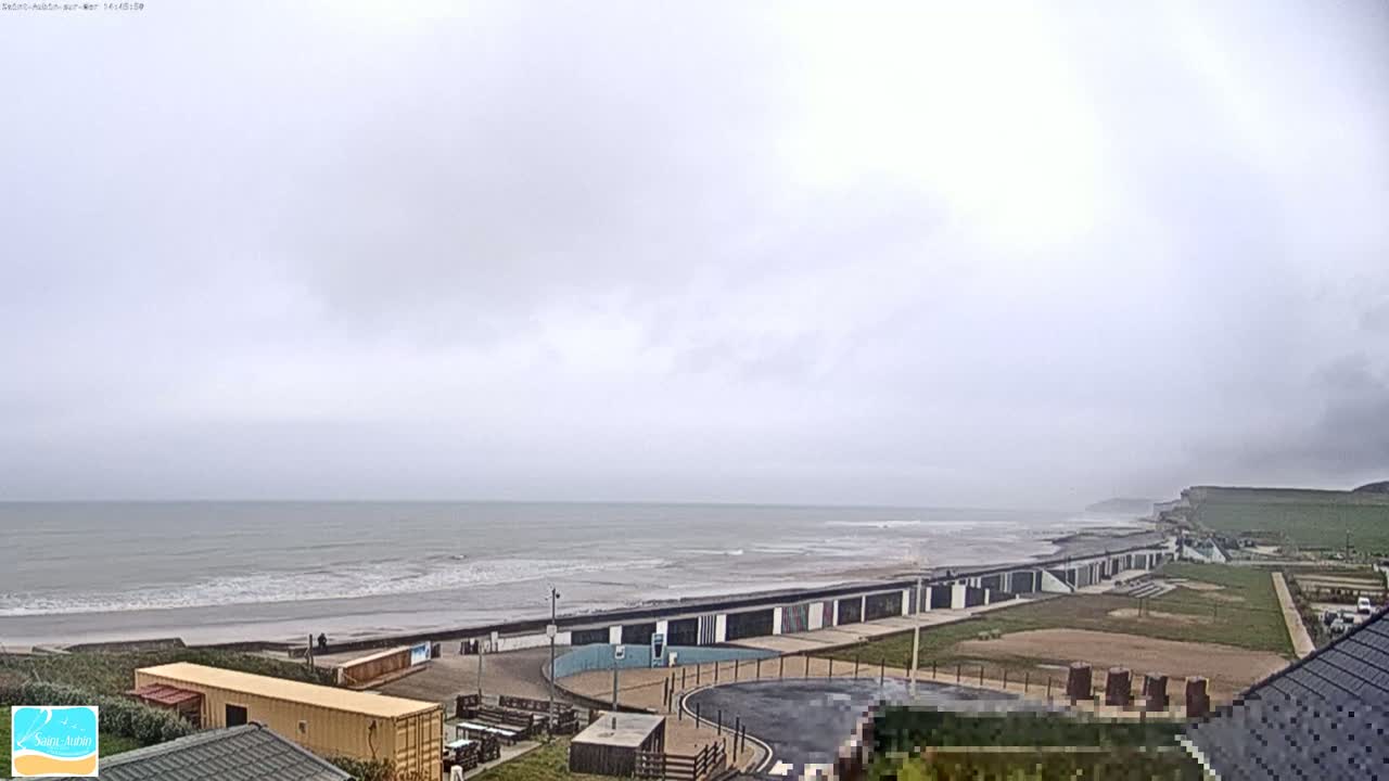 A gray, overcast day shows a beach with gentle waves, a shoreline with buildings, and grassy areas leading up to a cliff in the distance.