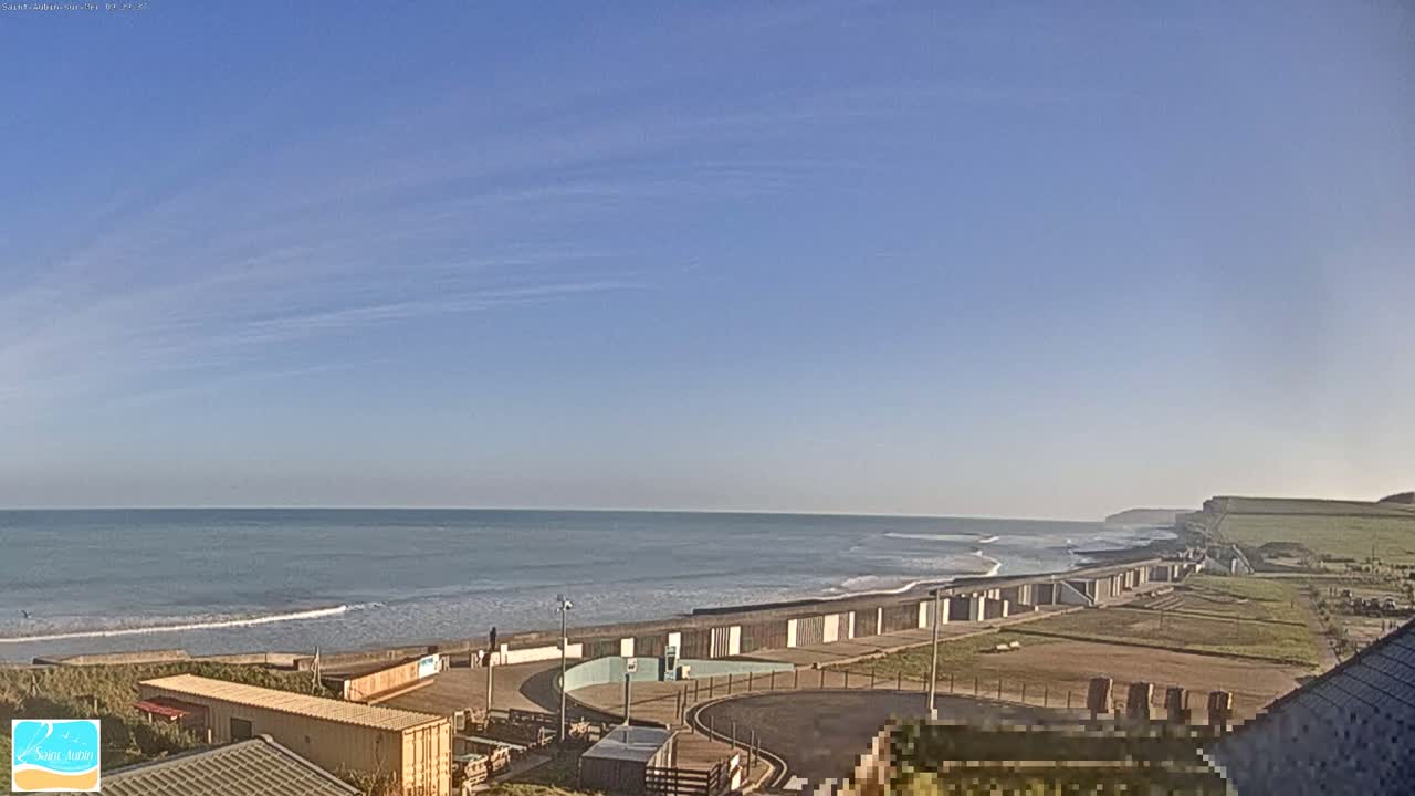 A mostly clear blue sky overlooks a calm ocean with small waves breaking on a beach, behind which sits a paved area with beach huts and some buildings.
