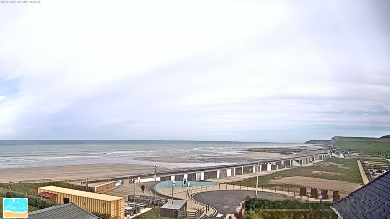 A mostly cloudy day shows a beach with calm waves, a seawall with small structures, and a paved area leading to buildings and cliffs in the distance.