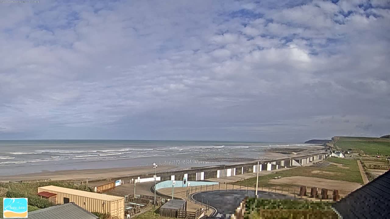 A beach with a seawall and small buildings is visible under a partly cloudy sky, with gentle waves rolling onto the shore.