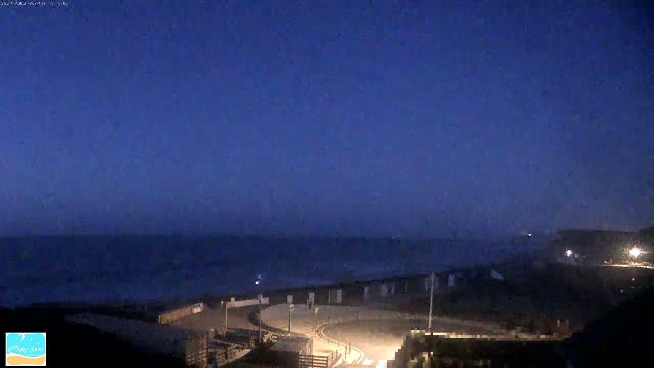 A dark blue nighttime sky overlooks a calm, dark ocean with a paved area and some buildings visible in the foreground.