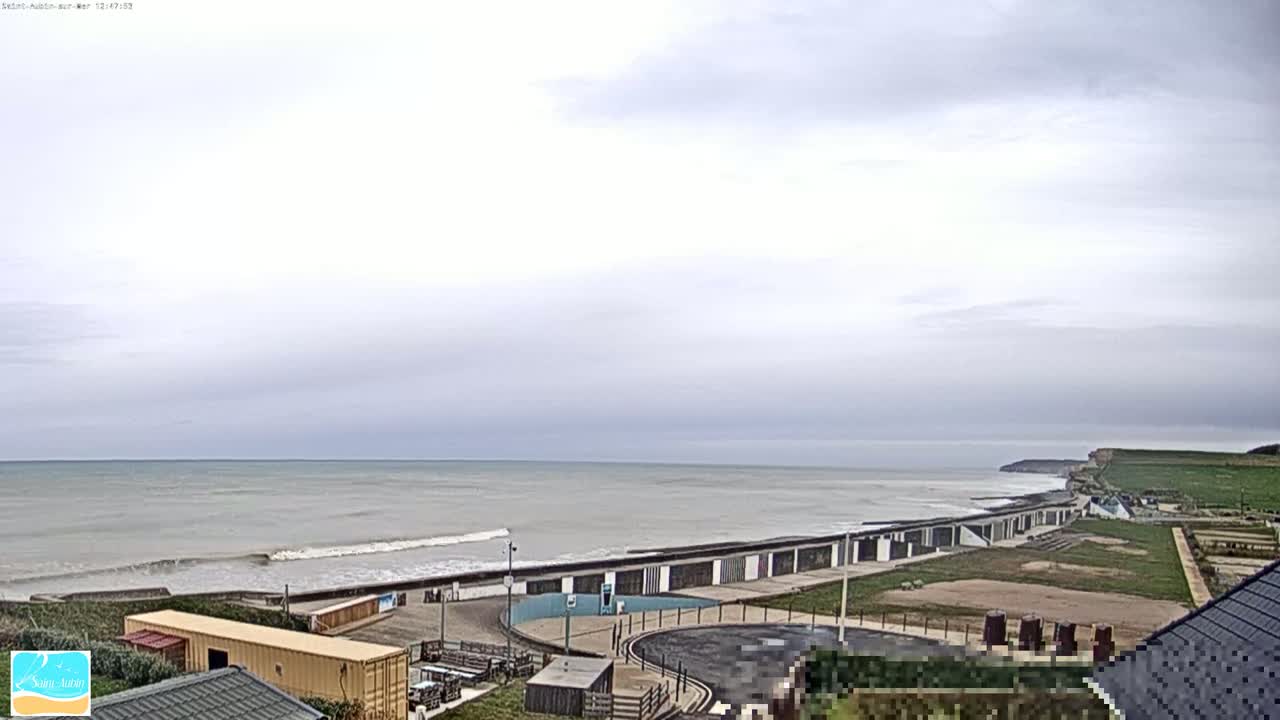 A mostly cloudy day shows a calm sea with small waves lapping a beach lined with buildings and a grassy area leading up to cliffs.
