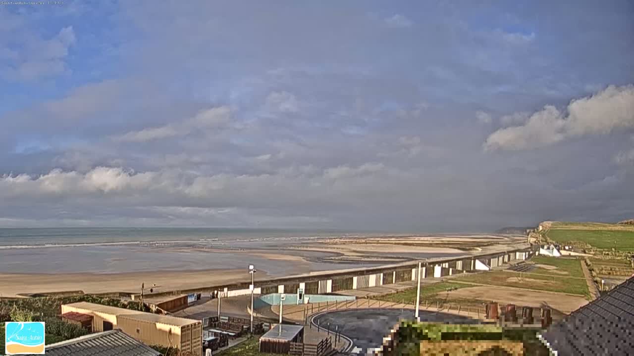 A mostly cloudy day reveals a wide sandy beach meeting a calm ocean, with a long seawall and other coastal buildings visible from a high vantage point.