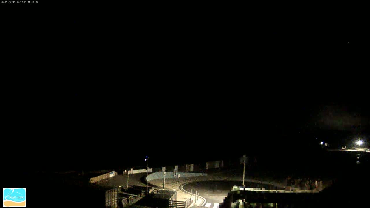 A nighttime, dark view of an empty paved area with faint lights and buildings in the distance under a mostly clear sky.