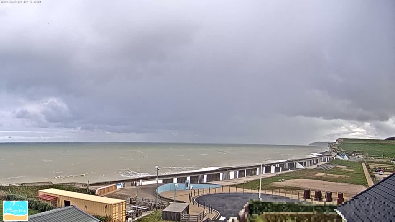 A mostly cloudy sky hangs over a gray sea meeting a coastline with buildings and grassy areas.