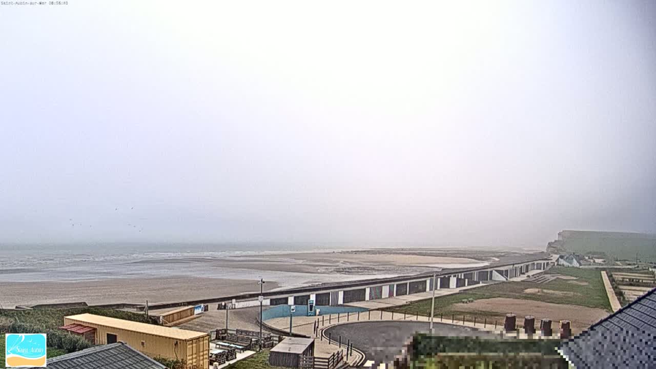 A foggy day reveals a long, low building complex near a wide, sandy beach and the ocean, with a cliff visible in the distance.