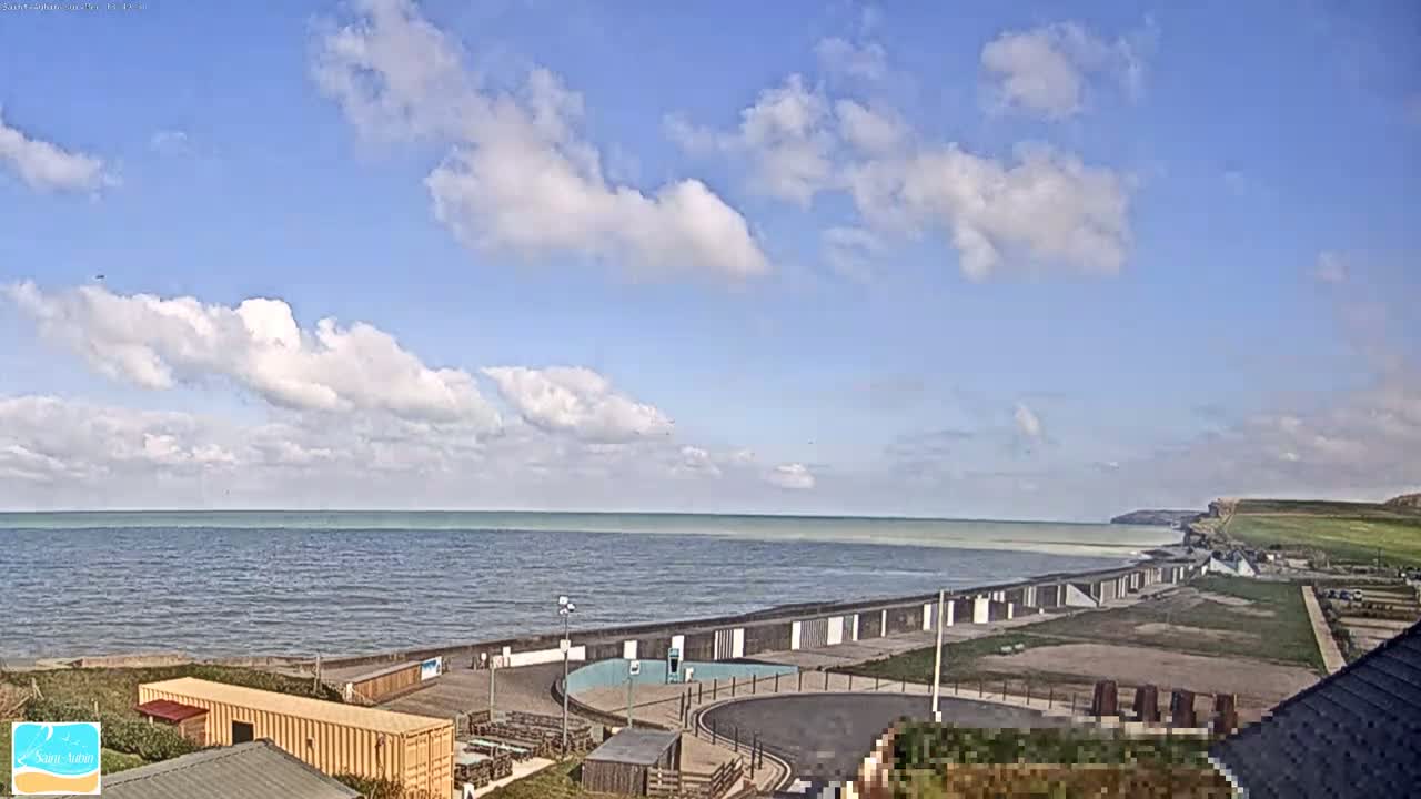 A mostly sunny day reveals a calm sea meeting a coastline with beach huts and a parking area, all under a partly cloudy sky.