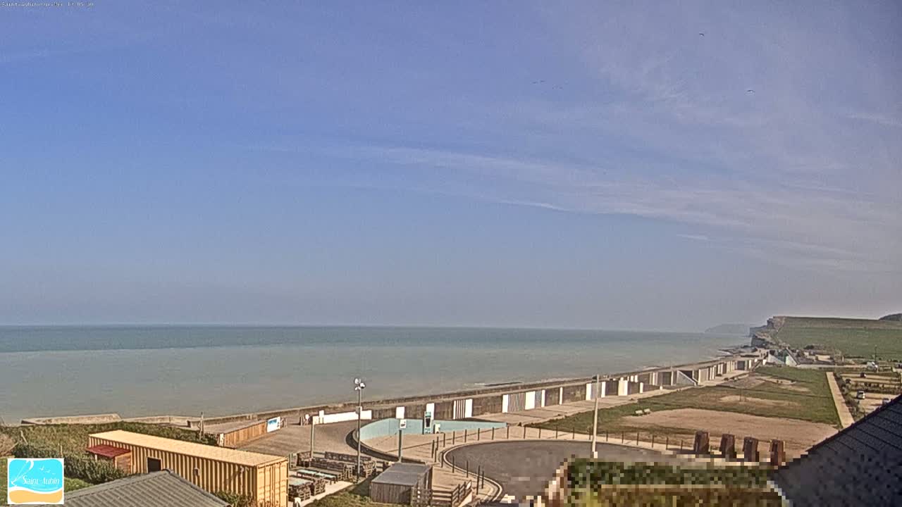 A calm, mostly clear day reveals a coastal scene with a wide, sandy beach, a seawall, and a parking area overlooking a tranquil ocean.