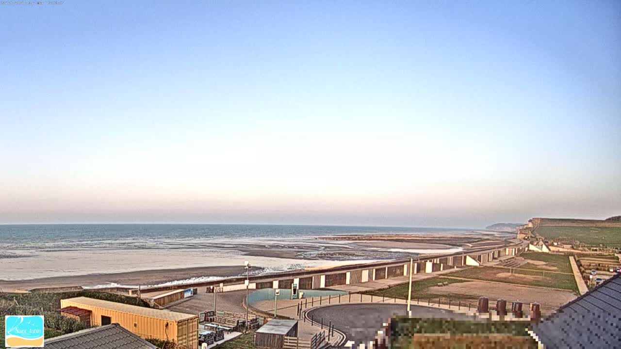A wide shot of a beach at low tide under a clear, light blue sky shows a row of beach huts and a coastal cliff in the distance.
