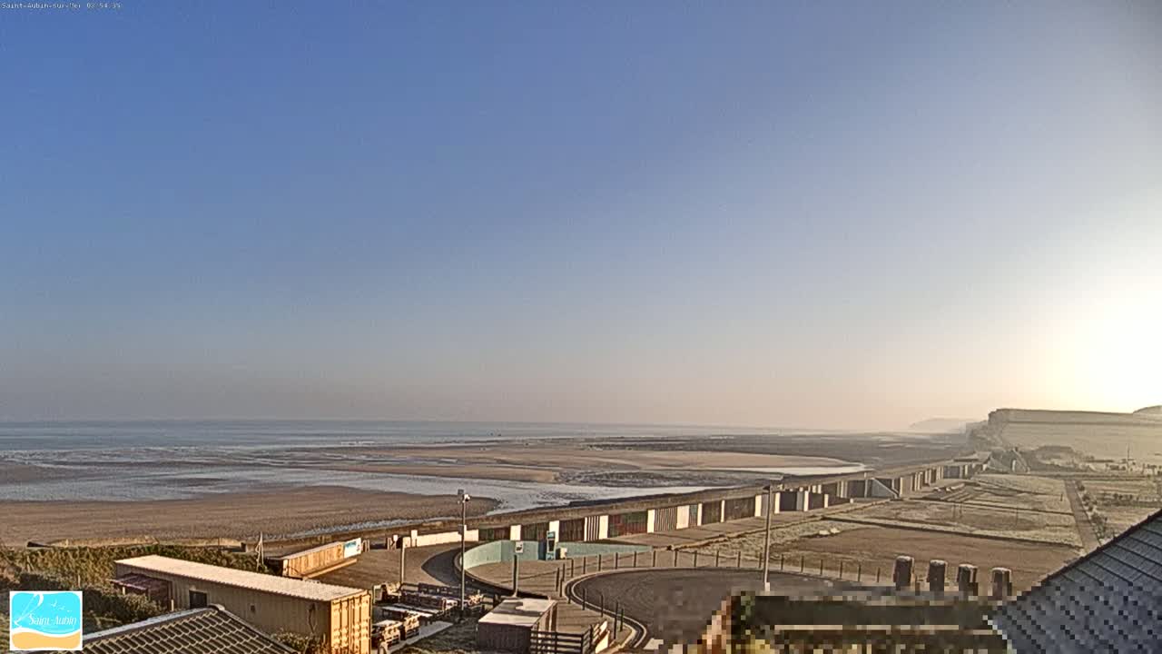 A wide, sandy beach stretches to a calm ocean under a clear, bright blue sky, with beach structures and buildings visible in the foreground.
