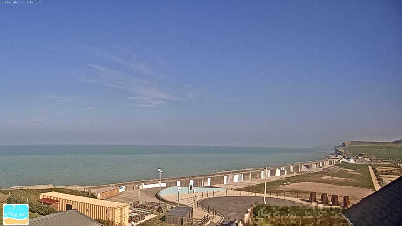 A calm sea stretches to the horizon under a mostly clear blue sky, with a sea wall and coastal buildings visible in the foreground.