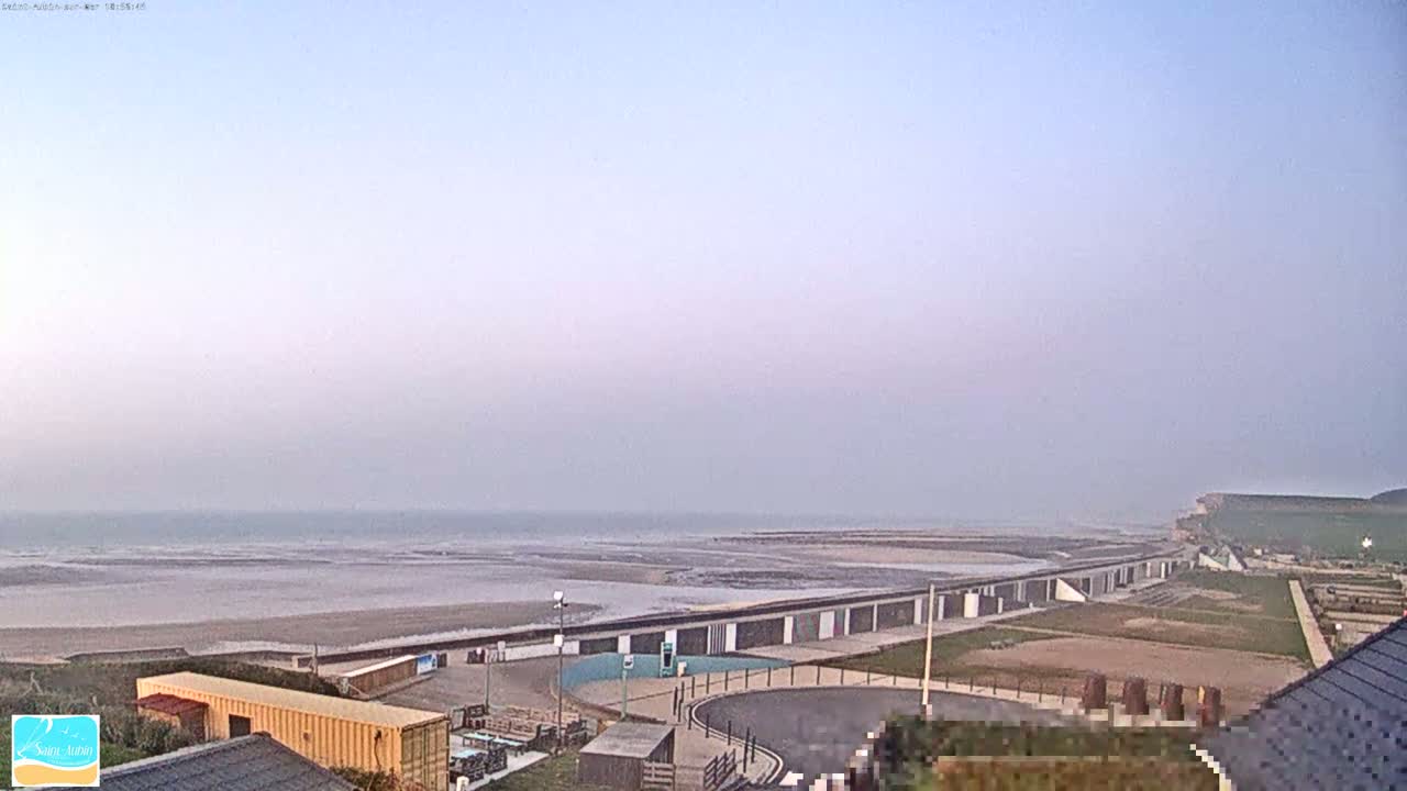 A hazy, overcast day reveals a beach with a long row of structures along the shoreline, and grassy areas leading to a cliff in the distance.