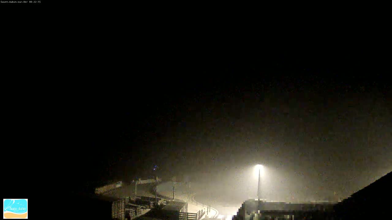 A foggy night scene shows a dimly lit road curving through a parking lot, illuminated by a single streetlight.