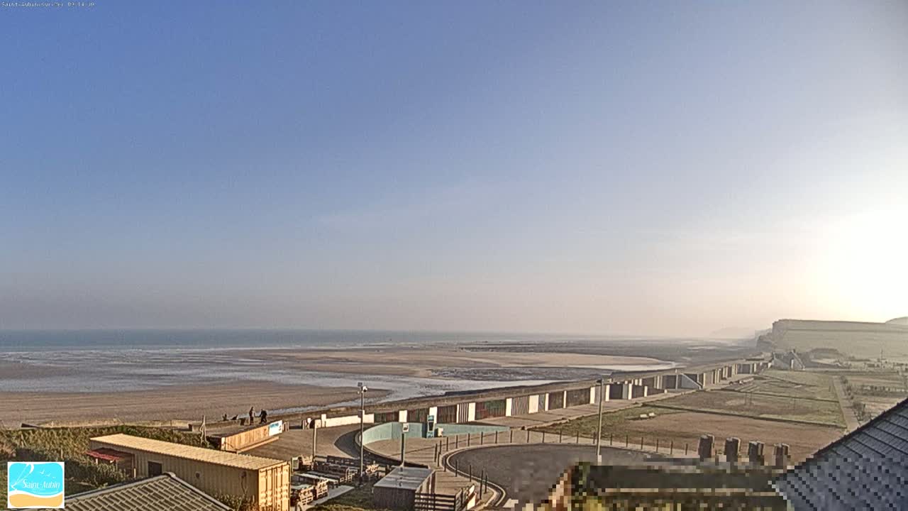 A beach with a calm ocean under a clear blue sky is visible from an elevated vantage point, showing beach huts and a paved area near the shore.
