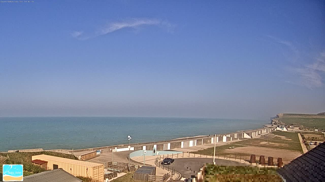A mostly clear blue sky overlooks a calm ocean meeting a seawall and grassy area next to a cliff on a sunny day.