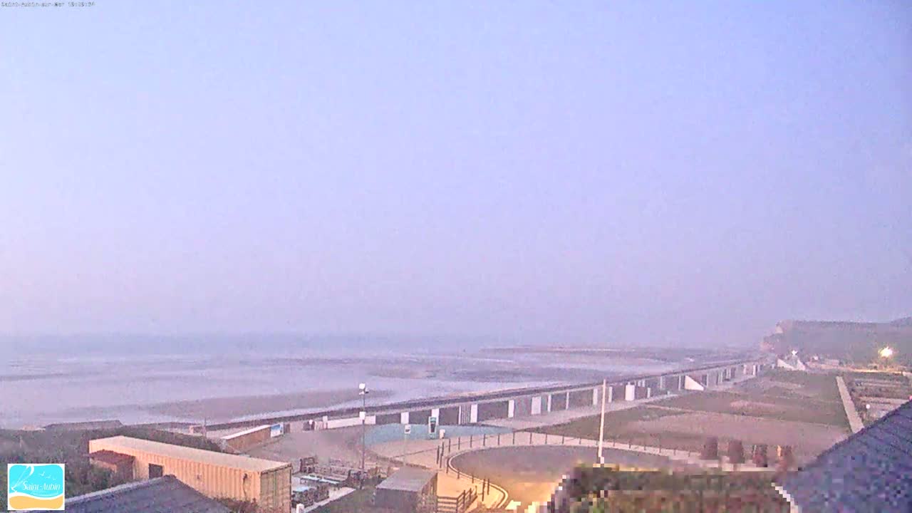 A hazy, pale-purple dawn illuminates a coastal scene featuring a long, low structure bordering a wide, flat beach that extends to a calm sea under a mostly clear sky.