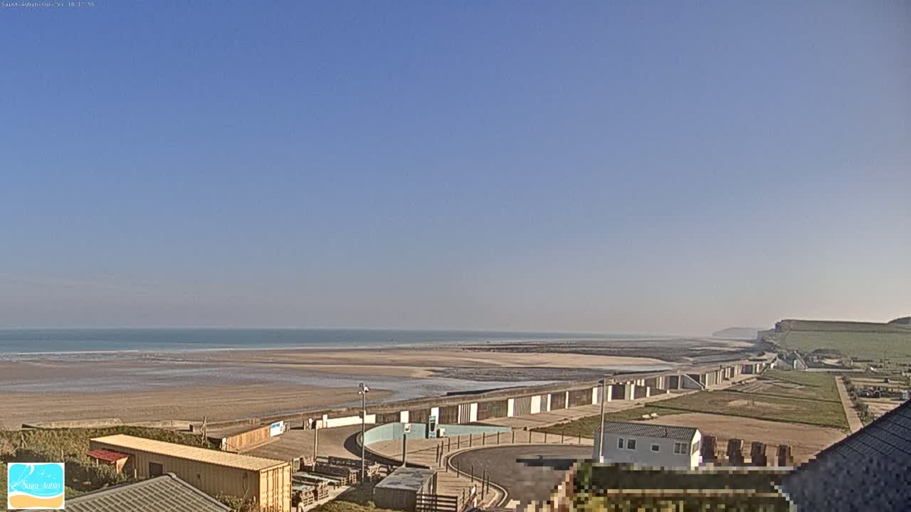 A wide sandy beach stretches to a calm ocean under a clear blue sky, with a seawall and buildings visible in the foreground.