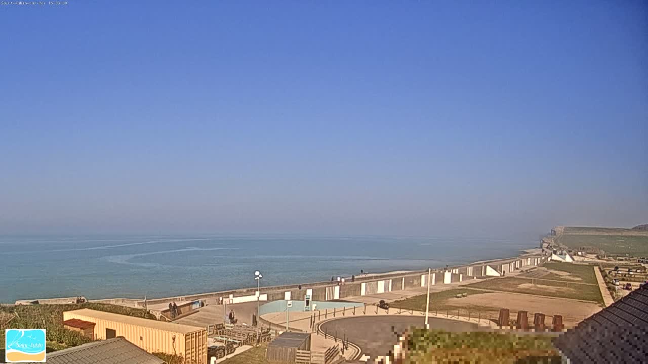 A calm ocean stretches to the horizon under a clear blue sky, with a coastal walkway and buildings visible in the foreground.