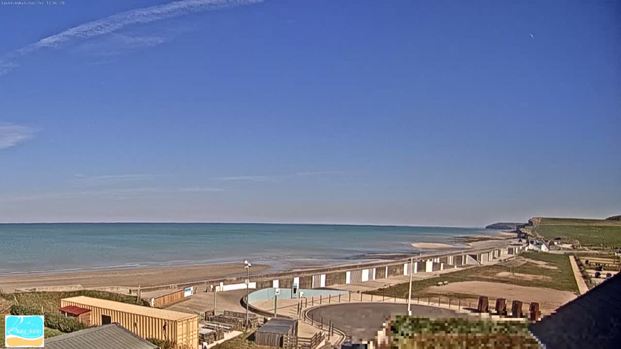 A sandy beach with calm, light-blue water stretches under a clear blue sky with a few wispy clouds, backed by a grassy cliff.