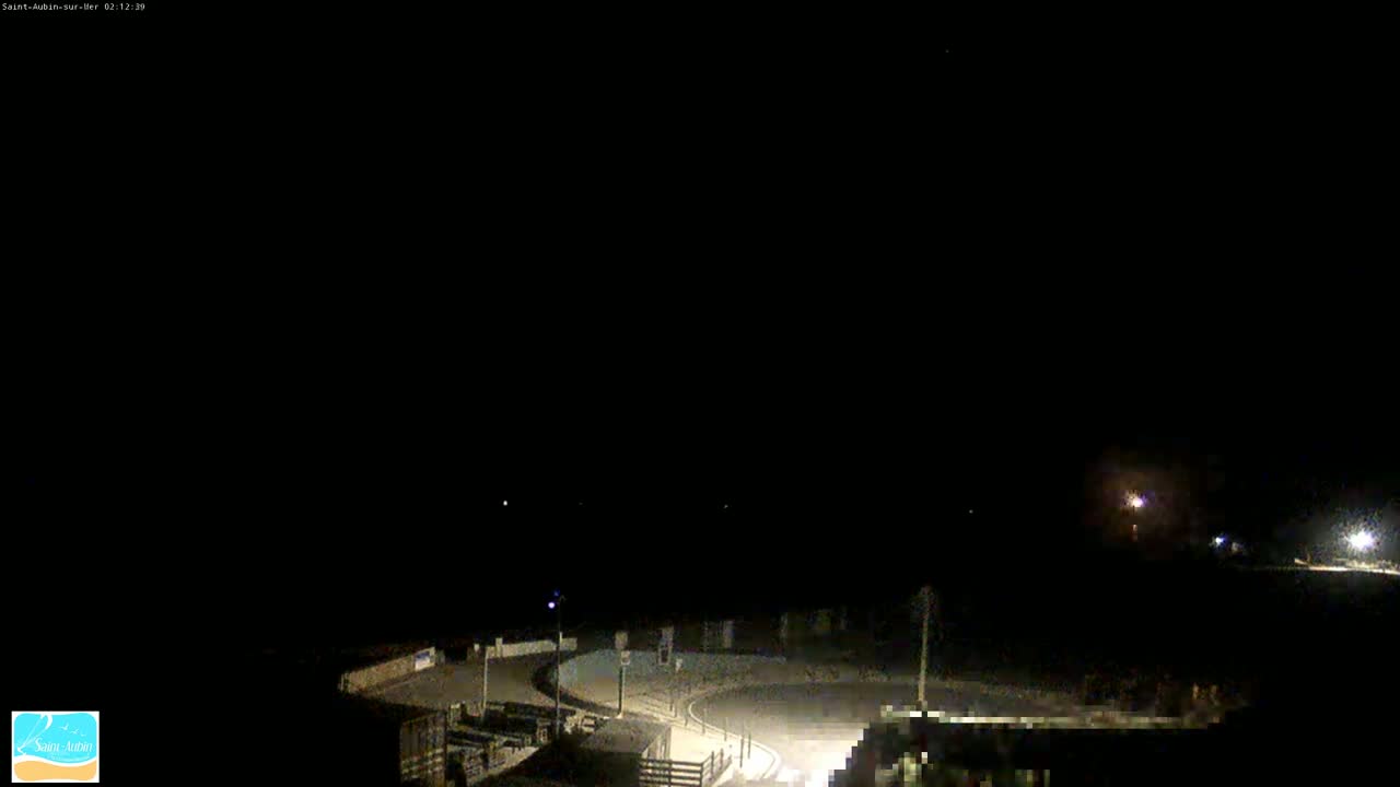 A nighttime, dimly lit outdoor scene shows a paved area curving around a small structure, with a few distant lights visible under a dark, clear sky.