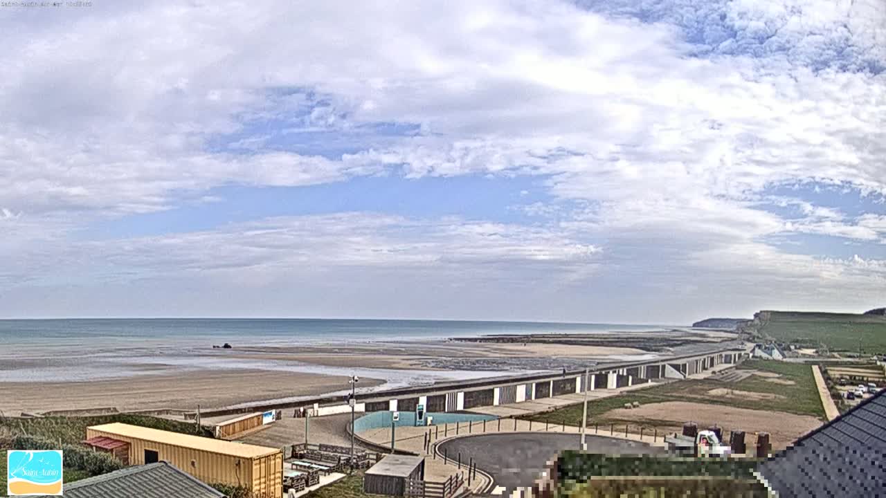 A partly cloudy sky overlooks a sandy beach, calm ocean, and a long row of beachside structures.