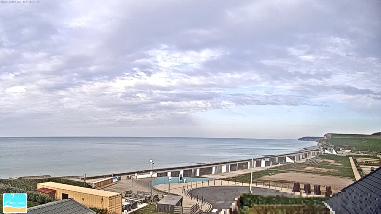 A calm ocean stretches to the horizon under a mostly cloudy sky, with a beach, seawall, and coastal buildings visible in the foreground.