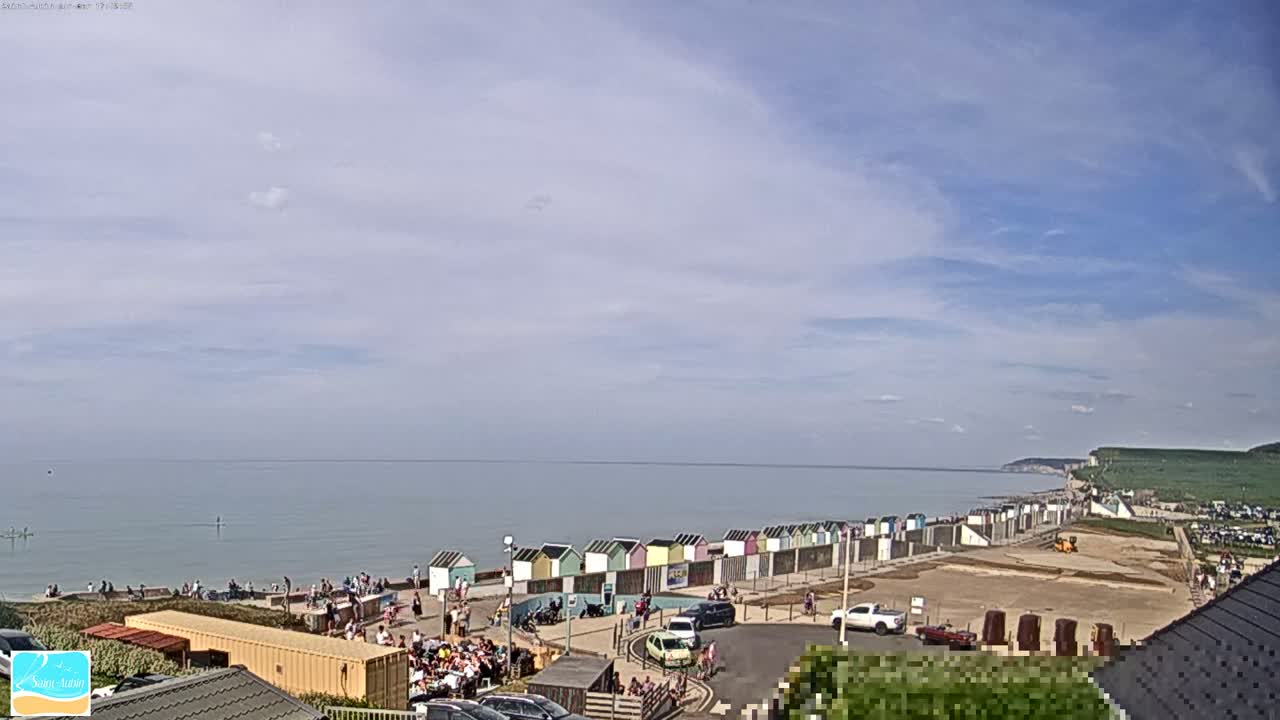 A mostly sunny day at a beach with calm water, featuring a row of colorful beach huts, people on the sand and promenade, and cars parked nearby, with a cliff visible in the distance.