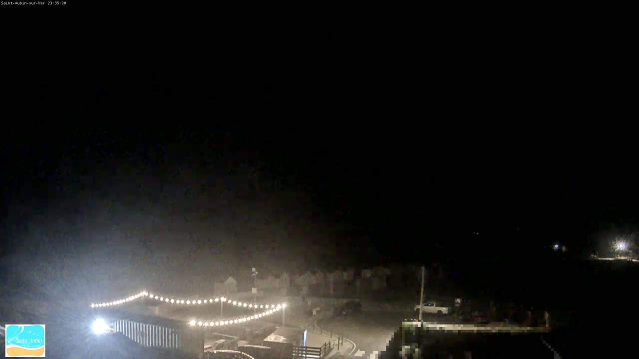 A nighttime, dimly lit outdoor scene shows a string of lights illuminating a small parking area with a few vehicles, some buildings, and a dark sky.
