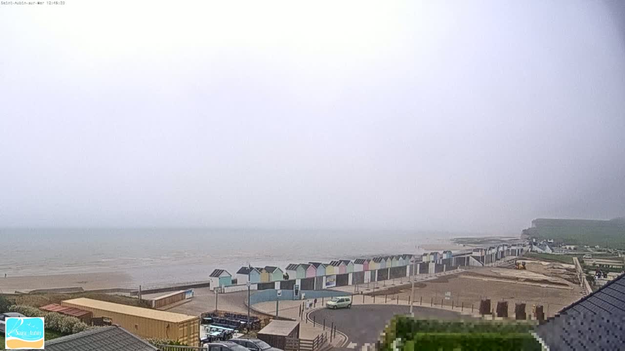 A hazy view shows a row of colorful beach huts along a sandy shore next to a calm sea, with construction taking place nearby.