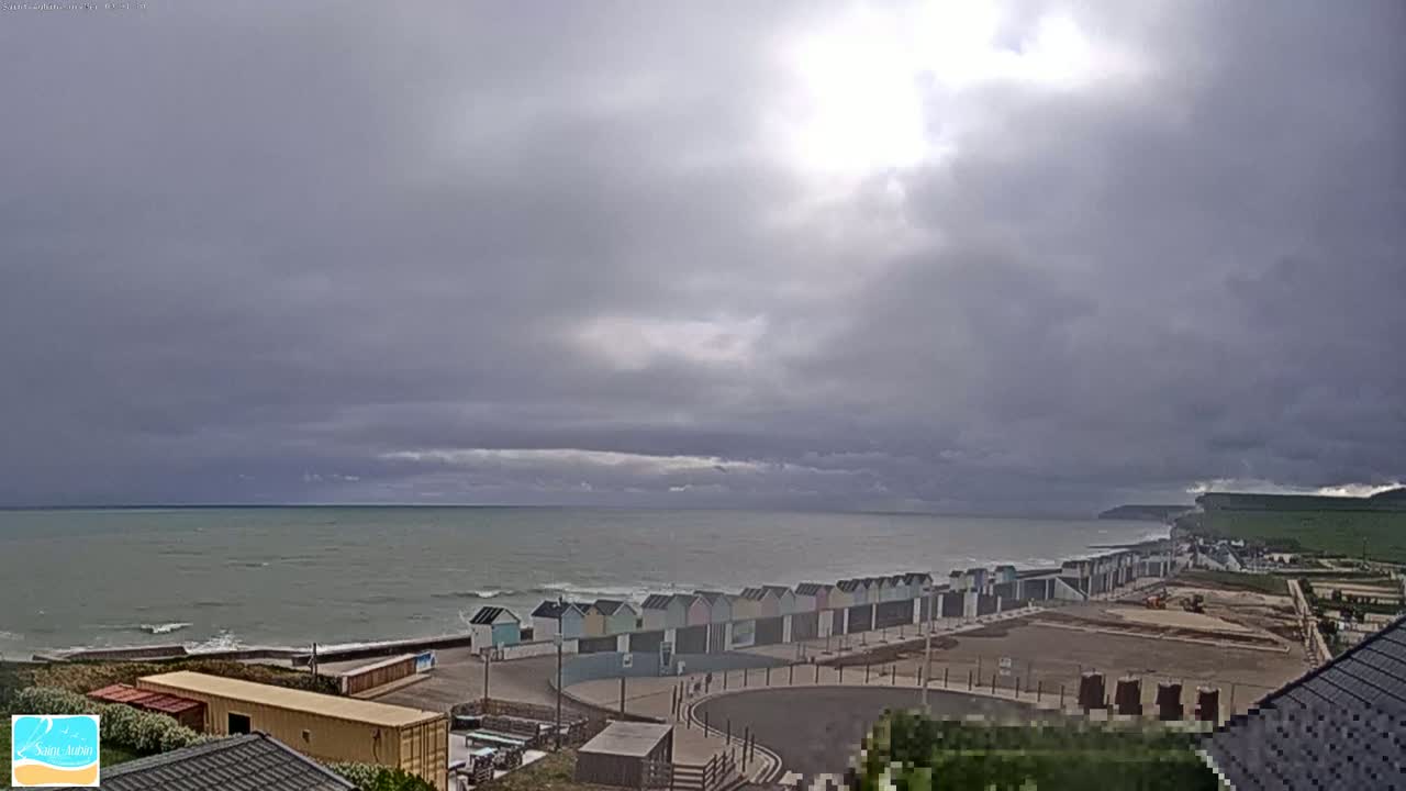 A row of colorful beach huts lines a coast under a mostly cloudy sky with the sun partially visible, overlooking a somewhat choppy sea.
